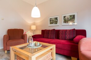 Un salon chaleureux avec un canapé rouge, une table basse en bois et une suspension. Deux tableaux encadrés ornent le mur.