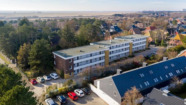 A multi-story building with a flat roof, surrounded by trees and parked cars.