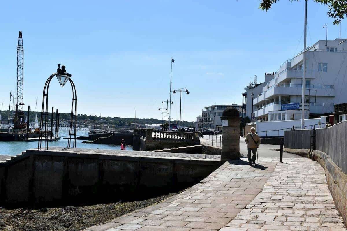 Eine Uferpromenade mit einem Pflasterweg, einer Metallbogenkonstruktion und einem Gebäude mit Balkonen.