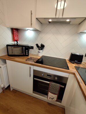 A compact kitchen with a microwave, toaster, and oven. There is a fire blanket on the wall and a container with kitchen utensils. - OLD SMIDDY APARTMENT, BALLOCH, LOCH LOMOND SLEEPS 3 IN FAMILY ROOM (Balloch)
