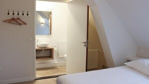A bedroom with a sloped ceiling, a bed, a small bathroom with a sink and mirror, and a door leading to another room.