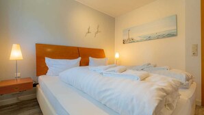A hotel room with a large bed, two bedside tables with lamps, a painting of a lighthouse, and a wall-mounted light switch. - Apartment "Flounder" (ID 344) - Friesenresidence (Sankt Peter-Ording)