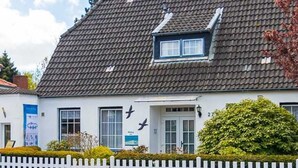 A white building with a grey tiled roof, a white picket fence, and a small garden area. - Apartment 4 (ID 036), Germany (Sankt Peter-Ording)