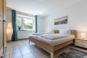 Un dormitorio con cama de madera, mesita de noche, lámpara, ventana con cortinas y un cuadro en la pared.