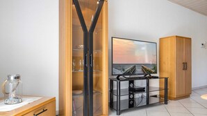 A living room with a wooden cabinet, a glass-fronted cabinet, a TV on a black stand, and a wooden cabinet with doors. - Apartment 4 (ID 036), Germany (Sankt Peter-Ording)