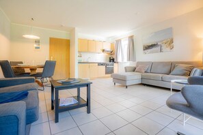 A modern living room with a grey sofa, a dining table with chairs, and a kitchen area with wooden cabinets and a stove. - Apartment 3B (ID 085), Germany (Sankt Peter-Ording)