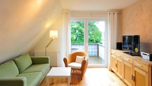 Un salon chaleureux avec un canapé vert, une table blanche, un fauteuil en osier, un téléviseur et un meuble en bois.