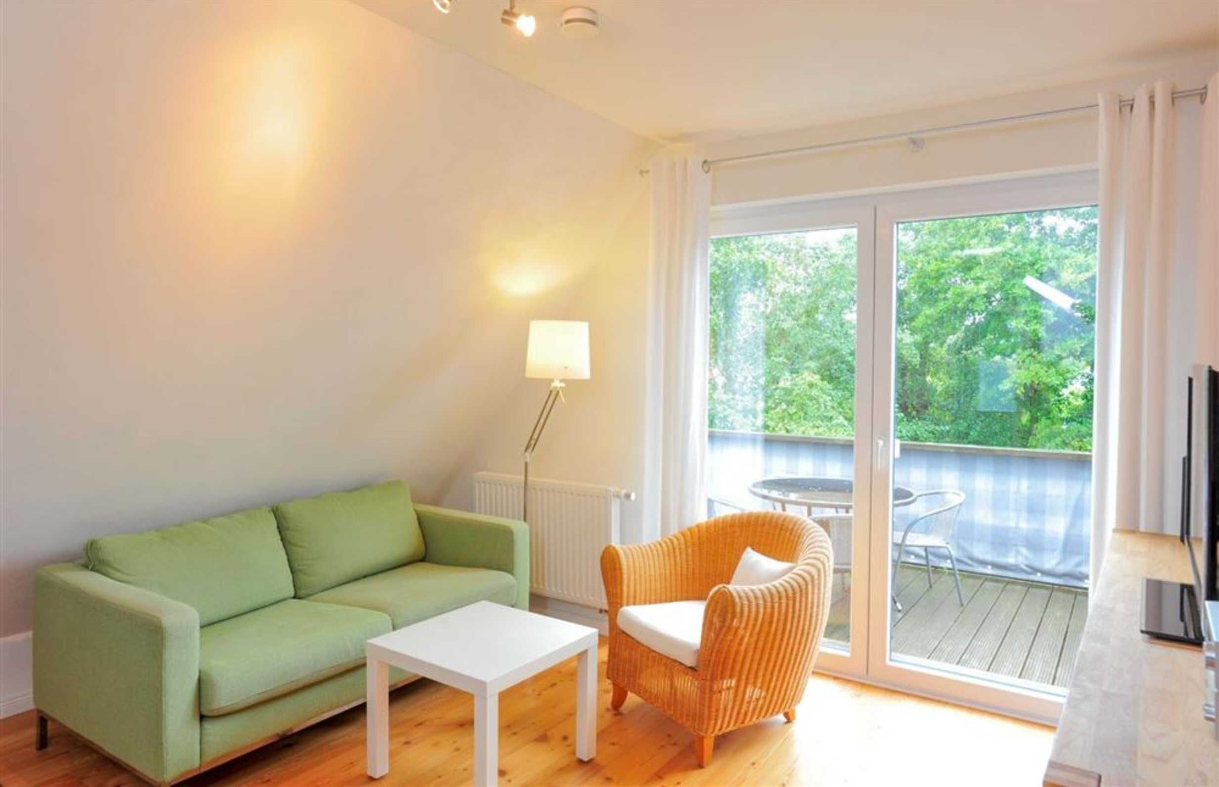 Un salón con un sofá verde, una mesa de centro blanca y una silla de mimbre. Hay un amplio ventanal corredero que da a un espacio exterior con una mesa y sillas.