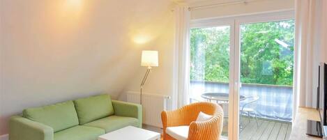 Un salón con un sofá verde, una mesa de centro blanca y una silla de mimbre. Hay un amplio ventanal corredero que da a un espacio exterior con una mesa y sillas.