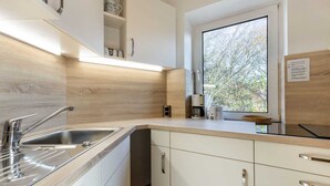 A modern kitchen with a built-in sink, white cabinets, and a window providing a view of trees. - Apartment 23 (ID 031), Germany (Sankt Peter-Ording)