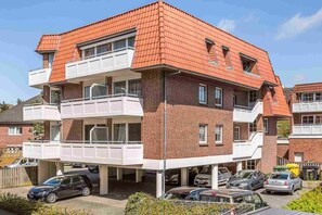 Un immeuble résidentiel à plusieurs étages, doté d’un toit recouvert de tuiles rouges, de deux balcons et d’un garage où sont stationnées plusieurs voitures.