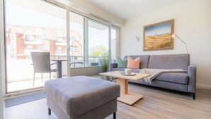 Un salon moderne avec un canapé gris, une table basse en bois et un pouf gris. Une grande fenêtre offre une vue sur les bâtiments à l’extérieur.