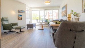Un salon moderne avec un canapé, des fauteuils et une table basse. De grandes fenêtres laissent entrer la lumière naturelle et offrent une vue sur l’extérieur.