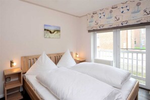 A bedroom with a wooden bed, white bedding, a nightstand with a lamp, a painting on the wall, and a window with a view of buildings. - Apartment 3 (ID 095), Germany (Sankt Peter-Ording)