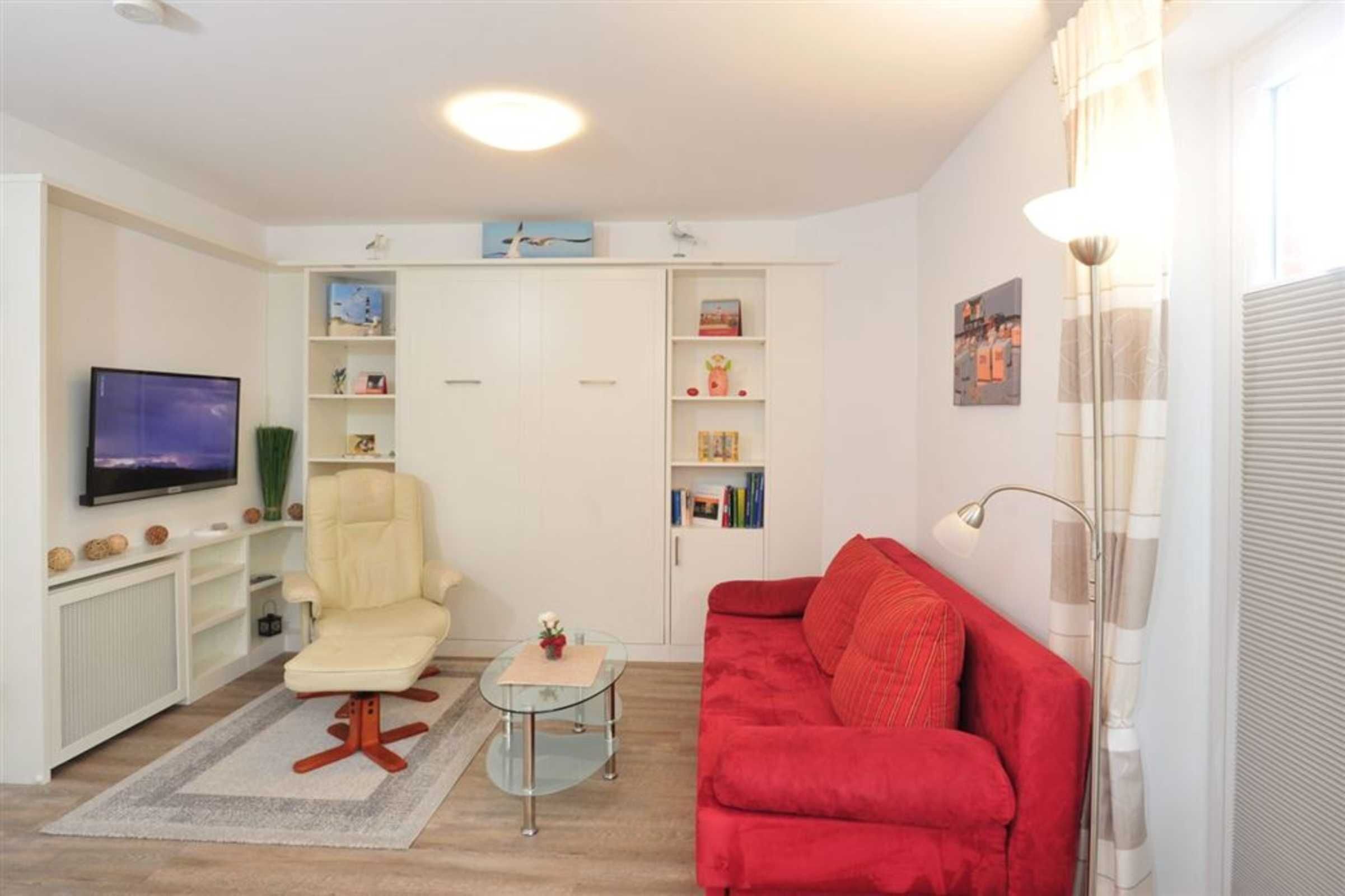 A living room with a TV, a red sofa, a cream armchair, a glass coffee table, and a bookshelf.