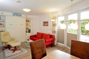 A modern living room with a red sofa, a beige armchair, and a dining table with chairs. There is a bookshelf, a floor lamp, and a window with curtains. - Apartment 3 (ID 095), Germany (Sankt Peter-Ording)