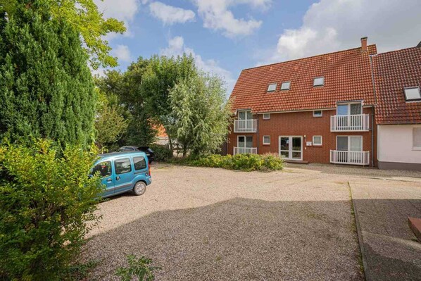 A two-story brick building with a red-tiled roof, a gravel driveway, and a parked blue car. - Apartment 3 (ID 095), Germany (Sankt Peter-Ording)