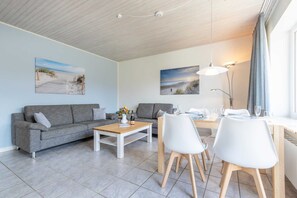 Uma sala de estar moderna com um sofá cinzento, uma mesa de centro de madeira, e um espaço de jantar com cadeiras brancas e uma mesa. Há duas pinturas nas paredes, um abajur e uma janela com cortinas.
