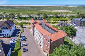 A multi-story building with a red-tiled roof, a parking lot with several cars, and a view of the sea in the background. - Apartment 12 (ID 232), Germany (Sankt Peter-Ording)