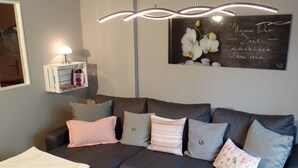 Ein modernes Wohnzimmer mit grauem Sofa, vielen Kissen, einer weißen Decke, einem kleinen Tisch mit Kerze und Blumen sowie einer Wandleuchte.