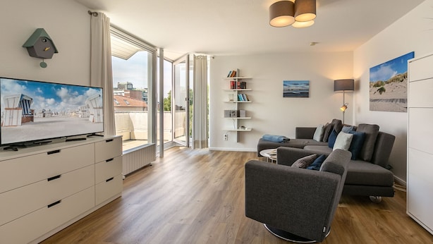 A modern living room with a flat-screen TV, a grey sofa, a bookshelf, and a large window with a city view.