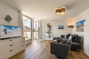 Uma sala de estar moderna com televisão de tela plana, um sofá cinza, uma estante de livros e uma janela grande com vista para a cidade.