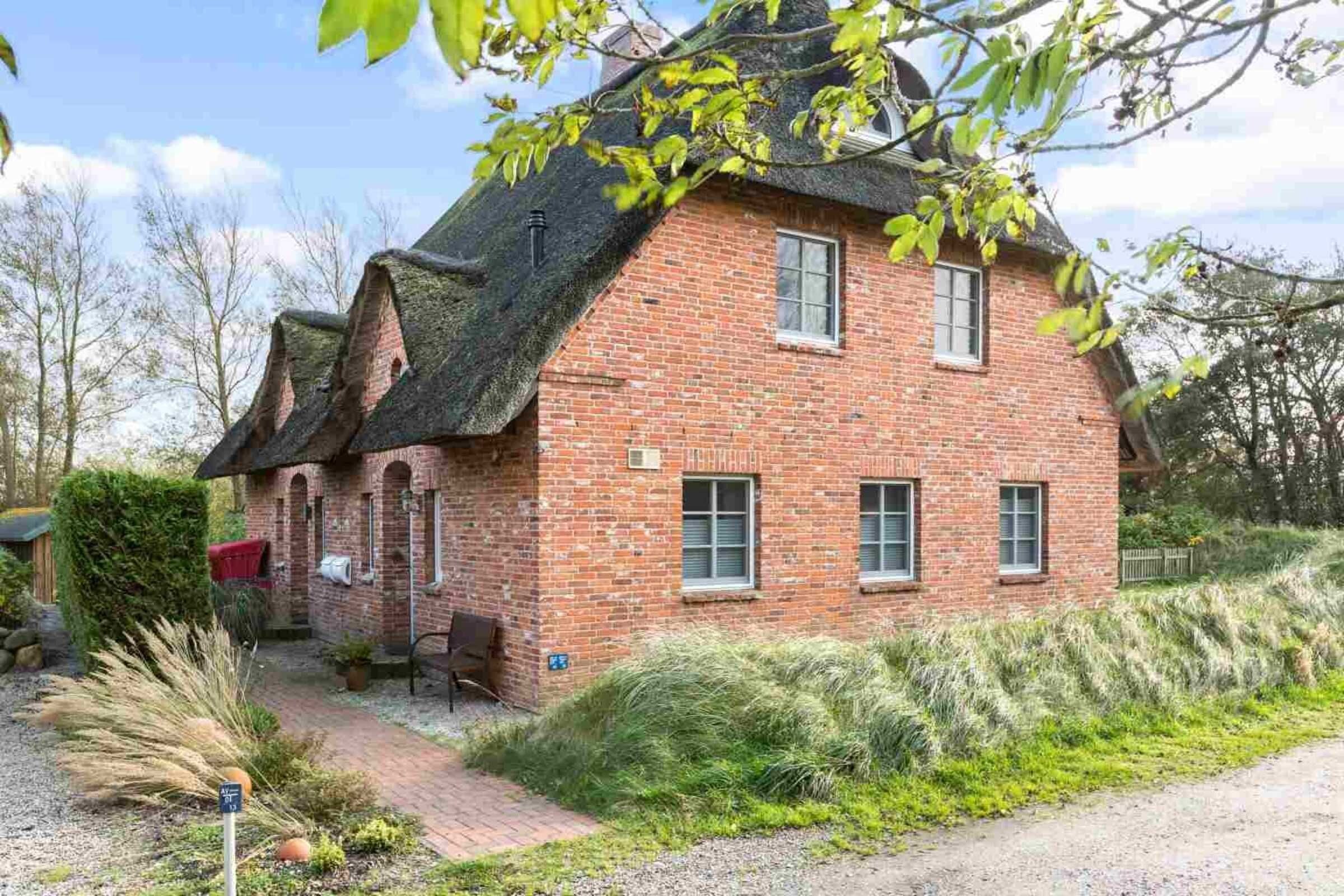 A brick house with a steeply pitched roof, a small porch, and a garden with various plants and a pathway leading to the entrance.