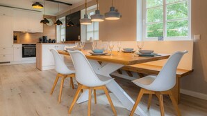 A modern kitchen with white cabinets, a wooden dining table, and white chairs. There are hanging pendant lights and a window with a view of trees.