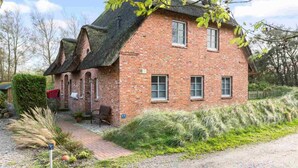 A brick house with a steeply pitched roof, a small porch, and a garden with various plants and a pathway leading to the entrance.