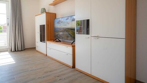 A modern room with a wooden floor, white cabinets, a flat-screen TV, and a large window with curtains. - Apartment 14 (ID 212), Germany (Sankt Peter-Ording)