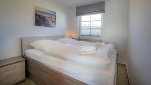 A neatly made bed with white linens and a painting of a beach scene on the wall. - Apartment 14 (ID 212), Germany (Sankt Peter-Ording)
