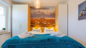 Ein Schlafzimmer mit einem Bett, einem Holzkopfteil mit Wandgemälde, weißen Schränken und einem Fenster mit Pflanze.