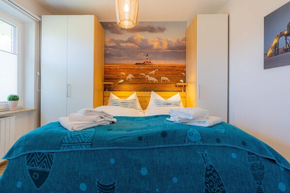 Ein Schlafzimmer mit einem Bett, einem Holzkopfteil mit Wandgemälde, weißen Schränken und einem Fenster mit Pflanze.