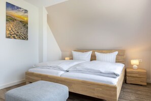 Un dormitorio con cama de madera, ropa de cama blanca, un banco gris y un cuadro de atardecer en la pared.