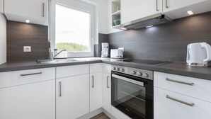 A modern kitchen with white cabinets, a built-in oven, a stovetop, and a window providing natural light. - Apartment OG (ID 135), Germany (Sankt Peter-Ording)