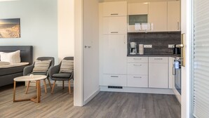 A modern kitchen with white cabinets, a black countertop, and a coffee machine. There is a gray sofa and two chairs with striped pillows in the adjacent living area. - Apartment ground floor behind (ID 136) (Sankt Peter-Ording)