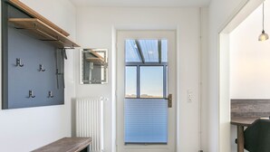 A modern entryway with a wooden bench, a mirror, a radiator, and a door with a view of the outdoors. - Apartment ground floor behind (ID 136) (Sankt Peter-Ording)