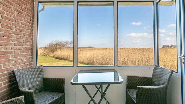 A room with a glass table and two wicker chairs, a brick wall, and a view of a field through large windows. - Apartment ground floor behind (ID 136) (Sankt Peter-Ording)