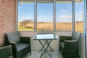 A room with a glass table and two wicker chairs, a brick wall, and a view of a field through large windows. - Apartment ground floor behind (ID 136) (Sankt Peter-Ording)