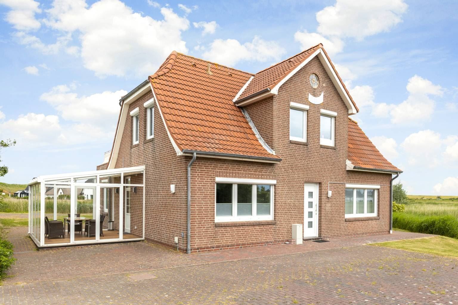 Une maison à deux étages, avec une façade en briques, un toit en tuiles et une véranda en verre aménagée avec du mobilier d’extérieur.