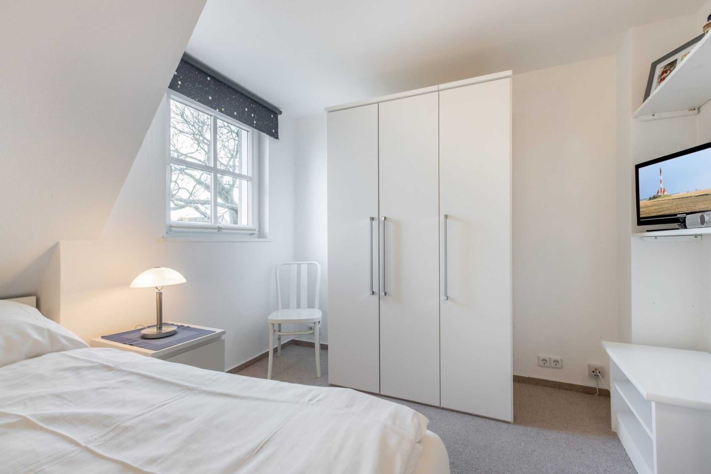 Une chambre à coucher comprenant un lit, une table de chevet avec une lampe, une chaise, une armoire et un téléviseur posé sur un meuble.