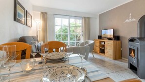 A dining area with a table set for four, a television, a fireplace, and a view of greenery outside. - House section 6 Port (ID 300) (Sankt Peter-Ording)