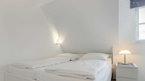 A neatly made bed with white bedding and pillows, a bedside lamp, and a small white nightstand. - House section 6 Port (ID 300) (Sankt Peter-Ording)