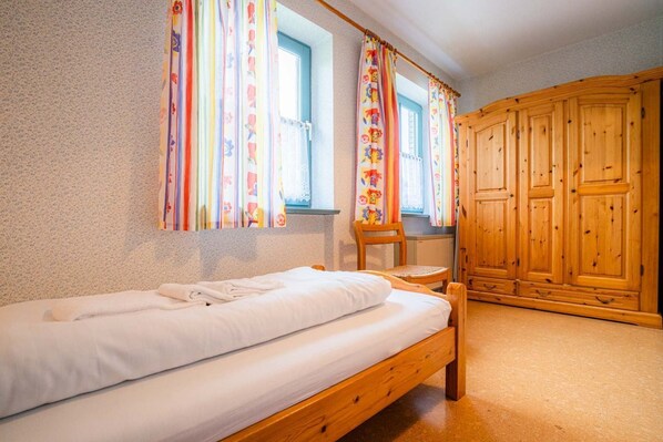 A single bed with a wooden frame, a chair, and a wooden wardrobe in a room with floral curtains. - Apartment OG (ID 313), Germany (Sankt Peter-Ording)