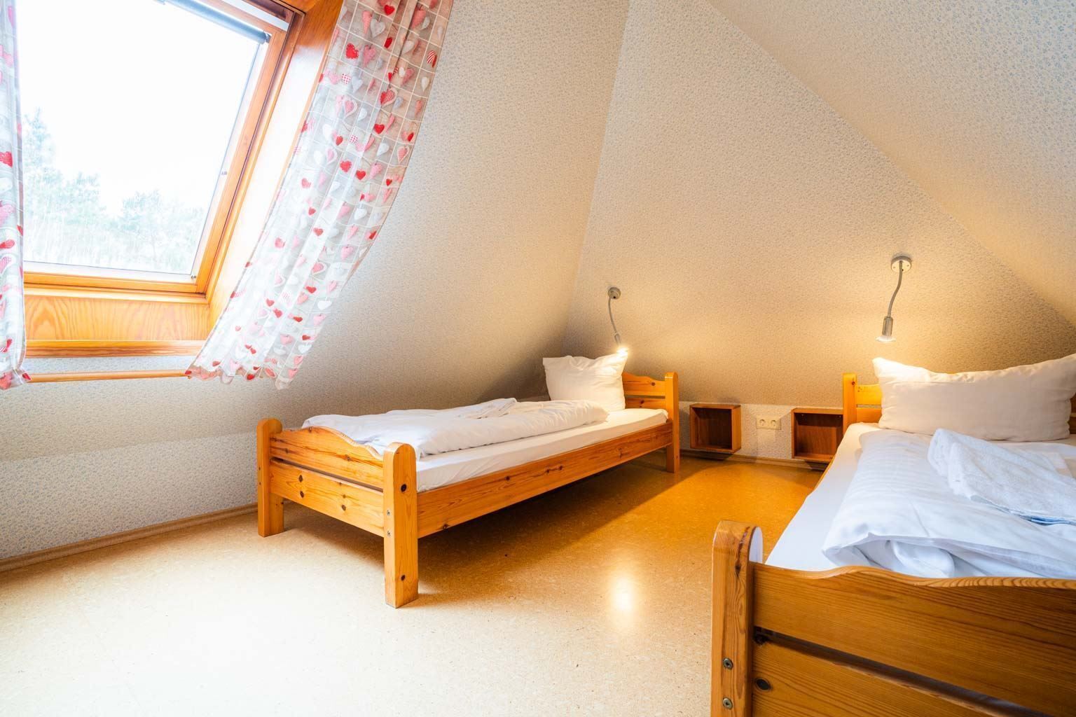 Ein kleines, gemütliches Schlafzimmer mit zwei Holzbett und einem Fenster mit Blumenvorhang, sowie einer geneigten Decke.