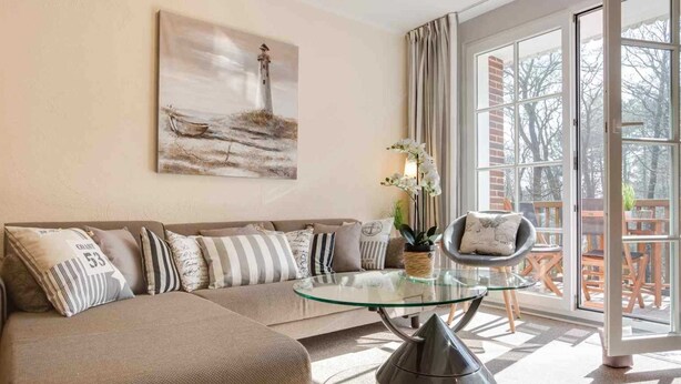 A modern living room with a glass coffee table, a beige sofa with striped cushions, a painting of a lighthouse, and a view through large windows.