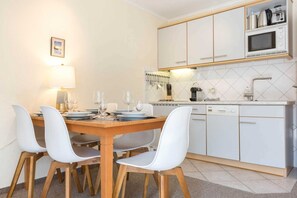 Une cuisine compacte avec des meubles blancs, un four à micro-ondes et un lave-vaisselle. On y trouve une table à manger avec des chaises et une lampe.