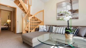 A modern living room with a glass coffee table, a sofa with striped cushions, a wooden staircase, and a painting of a lighthouse.
