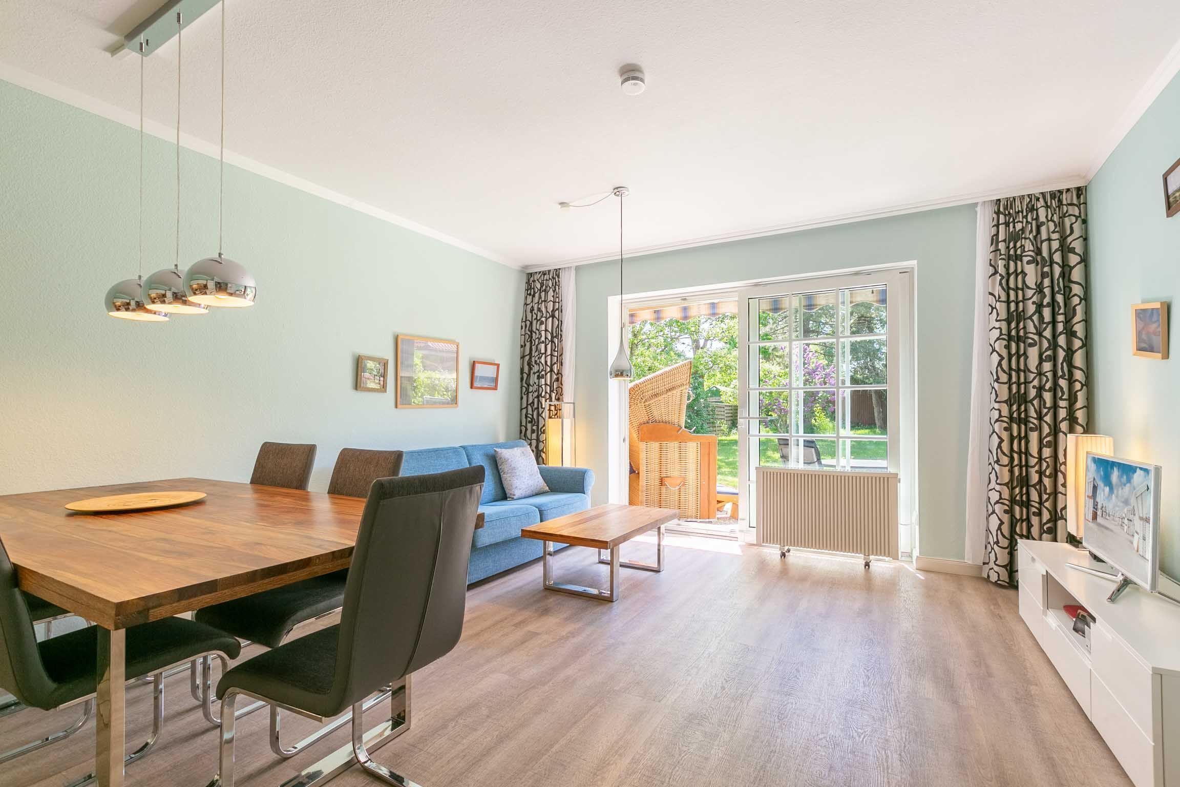 A spacious living room with a wooden dining table, comfortable seating, a sofa, and a large window with a view of greenery.