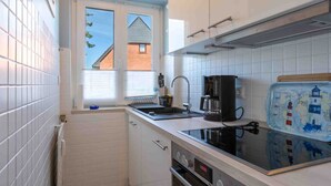 A compact kitchen with white cabinets, a black stove, and a window with a view of a house. - Apartment 1 "Starfish" (ID 292) (Room category) - Apartment 1 "Starfish" (ID 292) (Sankt Peter-Ording)
